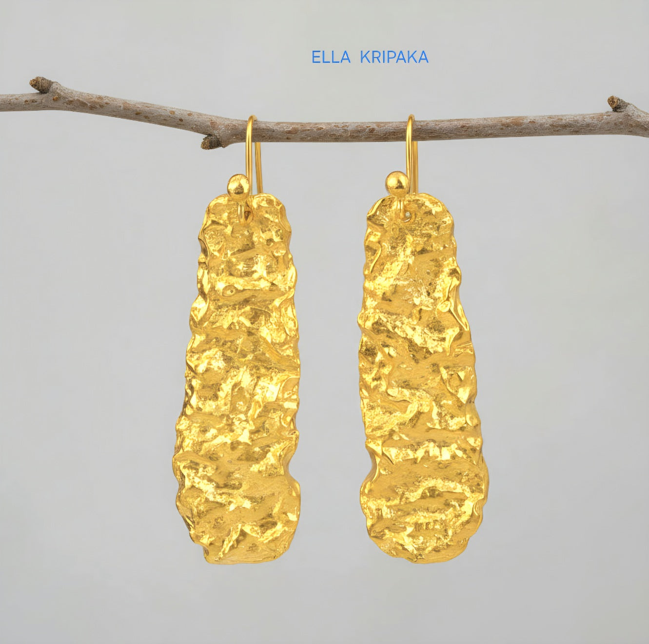 Organic hammered uneven 24k 9999 Gold earrings hanging on a branch and on a grey background, with 'ELLA KRIPAKA' branding