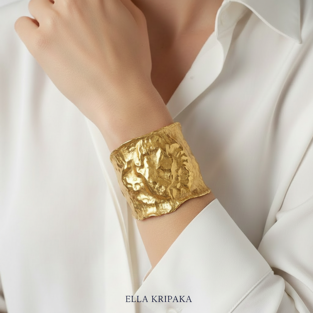 Hammered 24k gold 65mm cuff bracelet with convex uneven textures is worn by a woman on wrist and on white shirt background, and with artist 'Ella Kripaka' branding