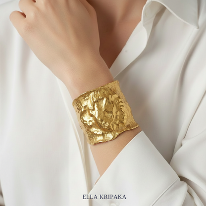 Hammered 24k gold 65mm cuff bracelet with convex uneven textures is worn by a woman on wrist and on white shirt background, and with artist 'Ella Kripaka' branding
