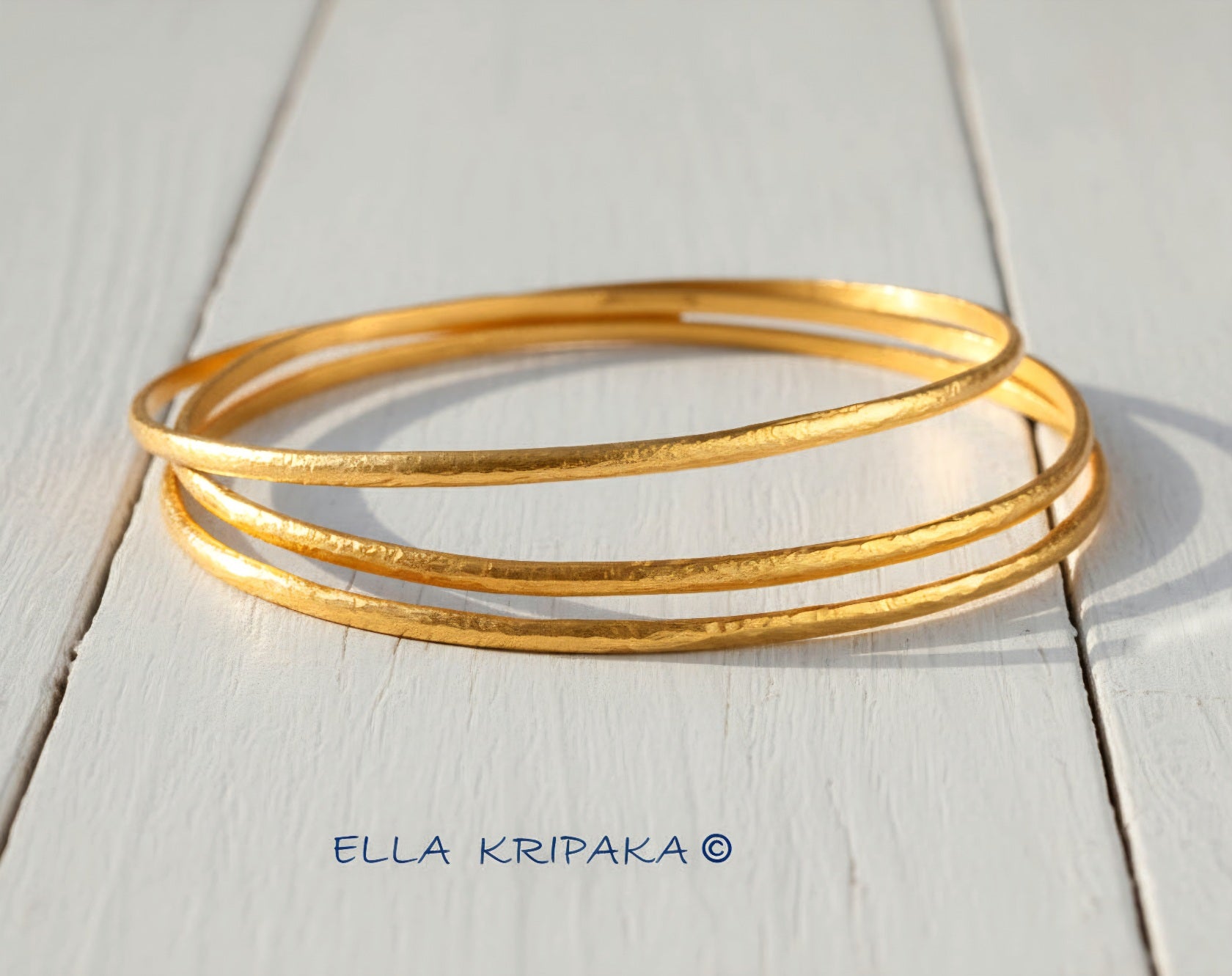 Hammered 22k gold three textured bangles bracelets Ancient Rome style on white wooden surface with 'Ella Kripaka' branding