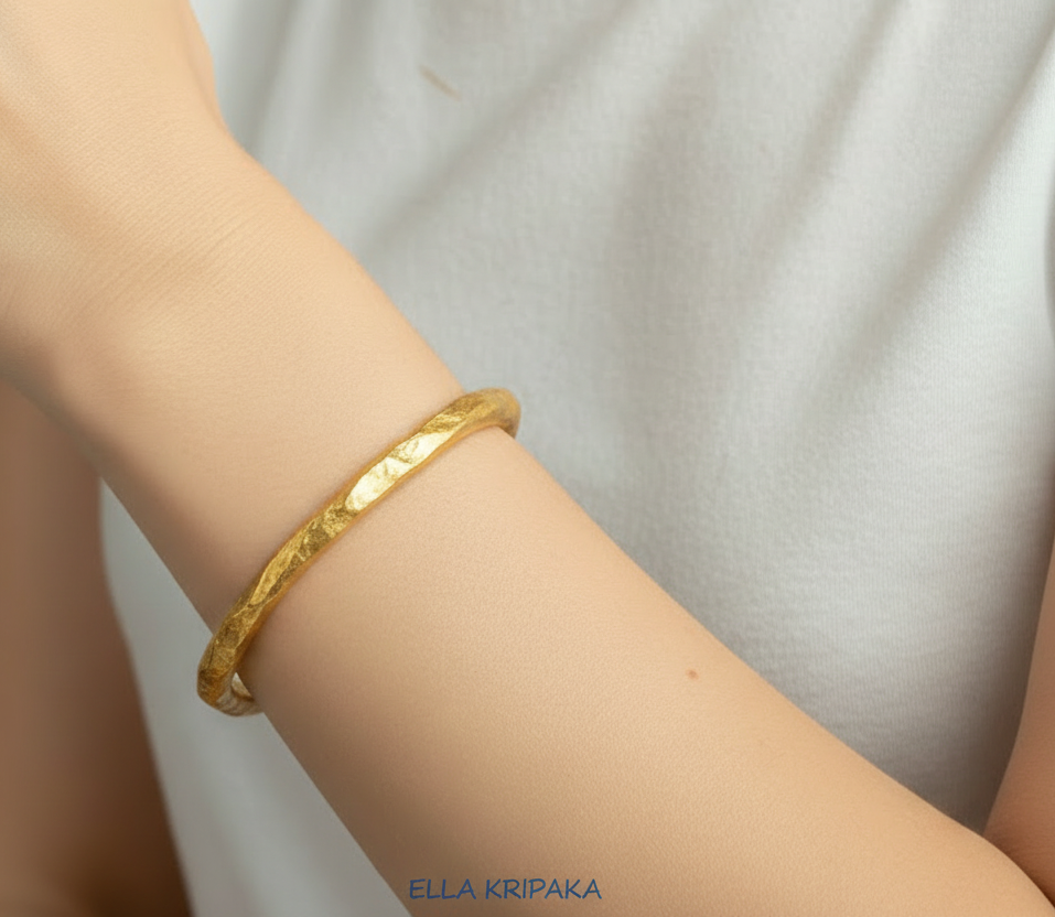 Organic hammered 24k 9999 Gold thick bangle uneven bracelet worn by a person on wrist and with white t-shirt background, artist's name 'Ella Kripaka' branding