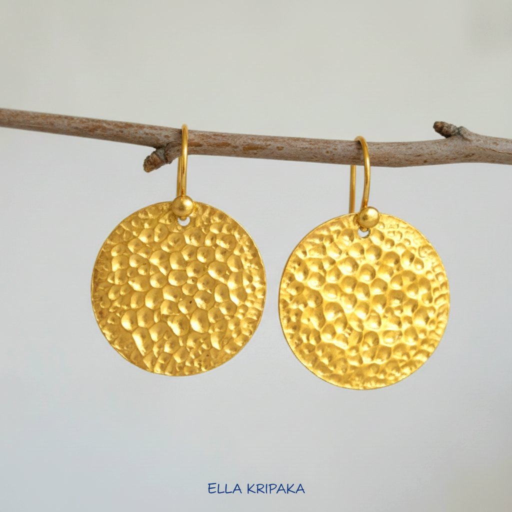 Organic hammered 24k gold, pure 9999 gold disk earrings with shiny forged gold textures hanging on a branch and with light grey background, artist-jeweler 'Ella Kripaka' branding