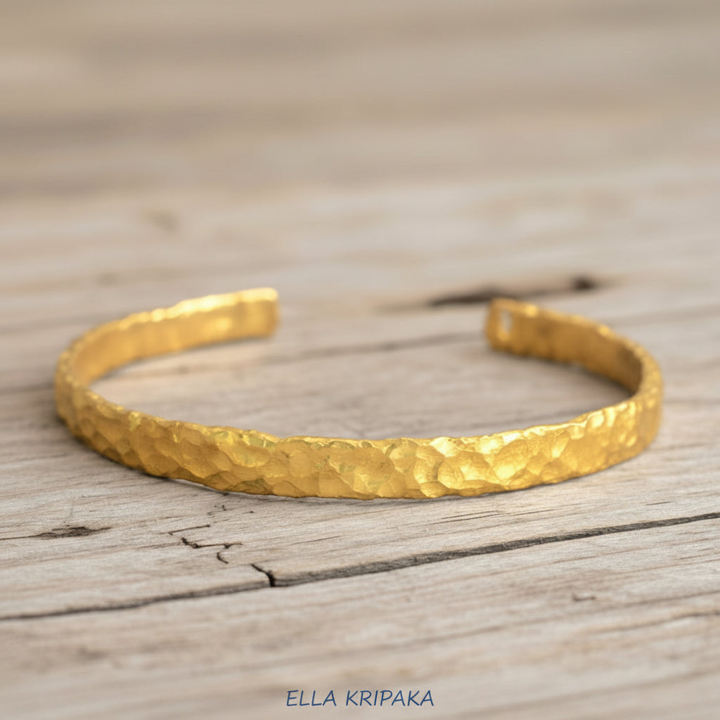 Organic hammered 24k gold bracelet cuff with uneven edges and textured design is on light wooden surface, branding artist 'Ella Kripaka'