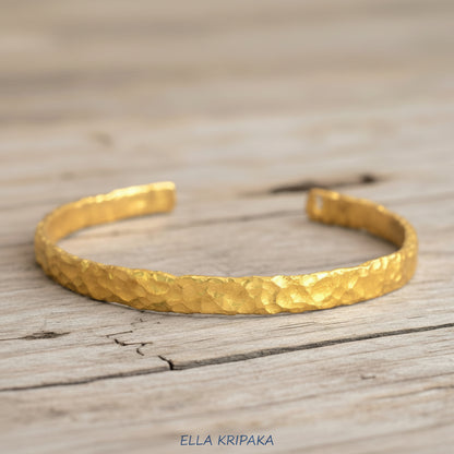 Organic hammered 24k gold bracelet cuff with uneven edges and textured design is on light wooden surface, branding artist 'Ella Kripaka'
