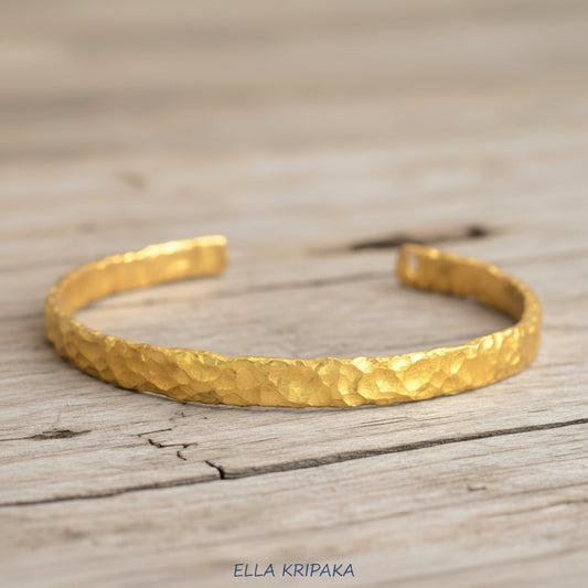 Organic hammered 24k gold bracelet cuff with uneven edges and textured design is on light wooden surface, branding artist 'Ella Kripaka'