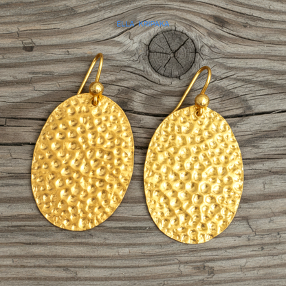 Organic 24k 9999 gold hammered oval earrings with shiny gold textures are on old wooden background, artist-jeweler's name 'Ella Kripaka'