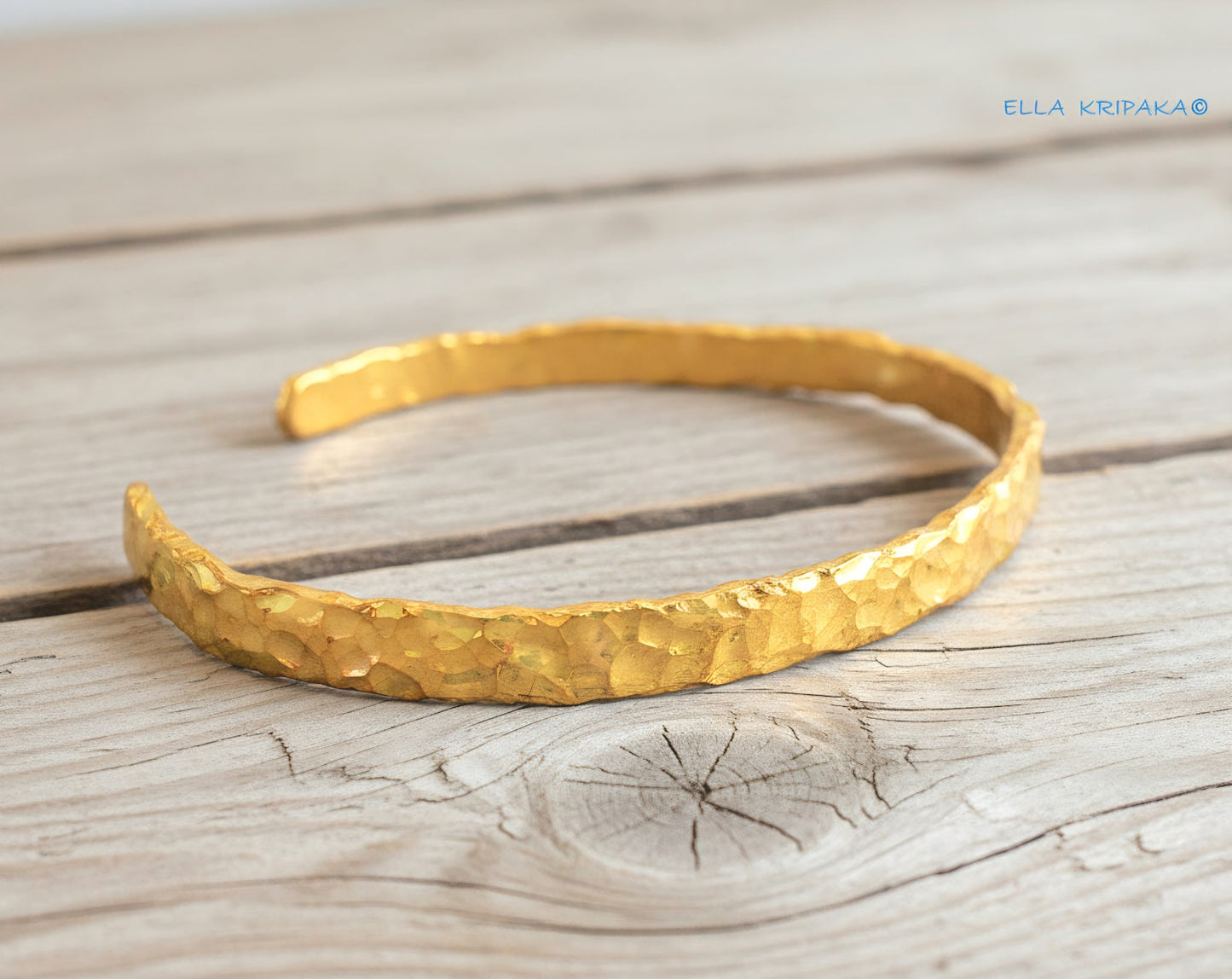 Organic hammered 24k gold bracelet cuff with thick uneven edges and textured design is on light wooden surface, branding artist's name 'Ella Kripaka'