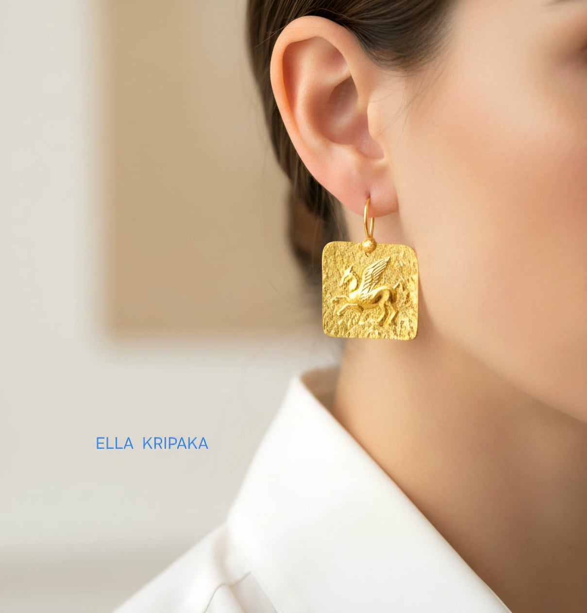 Hammered 24k 9999 gold square earring with convex Pegasus worn by a person with a blurred background, and artist's name 'Ella Kripaka'