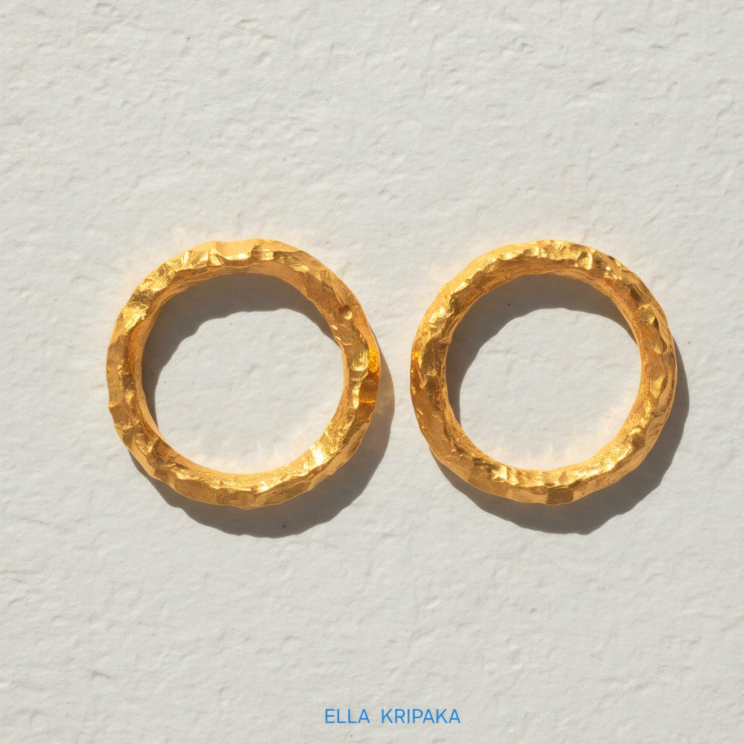 Two organic 24k gold hammered rings with uneven textured surface on a light grey craft paper background, branded 'Ella Kripaka'