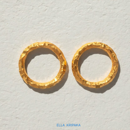 Two organic 24k gold hammered rings with uneven textured surface on a light grey craft paper background, branded 'Ella Kripaka'