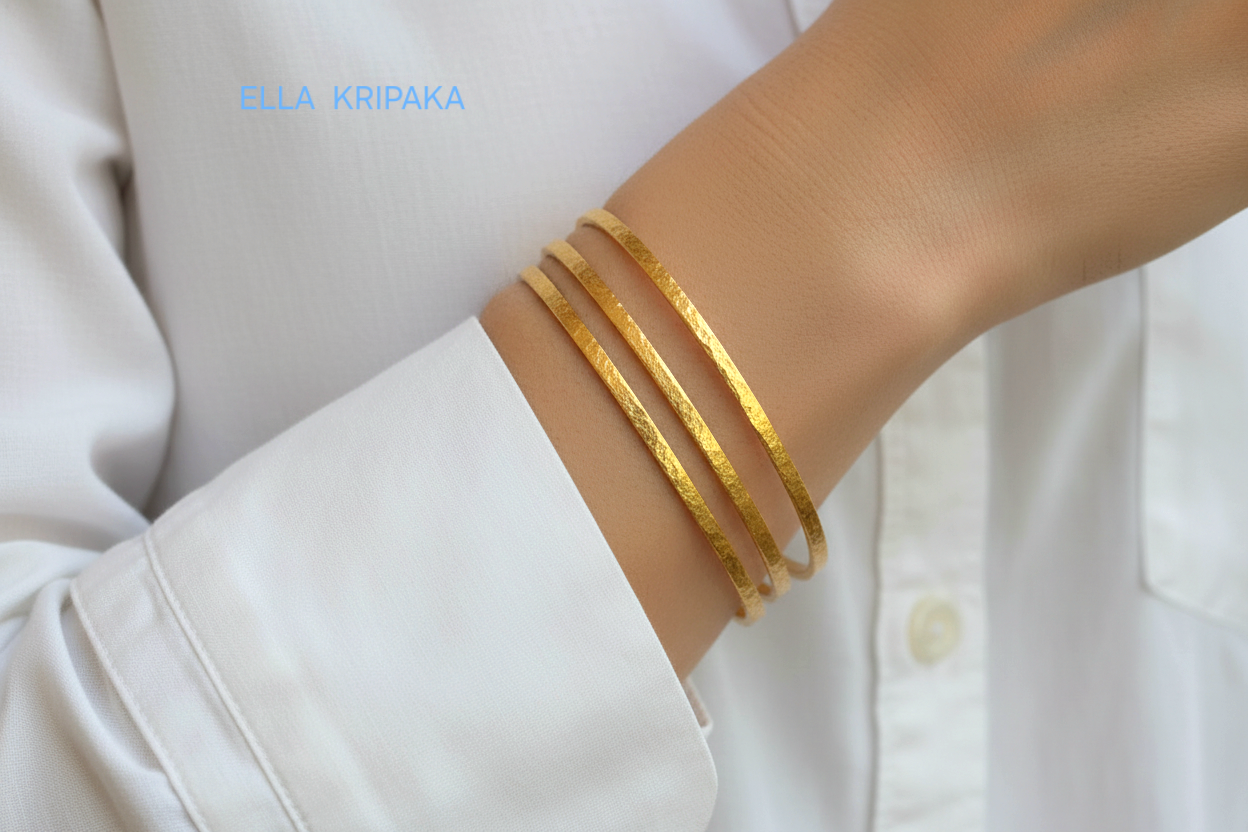 Hammered 22k gold three textured bangles bracelets Ancient Rome style on model's wrist and on white blurred shirt background, with 'Ella Kripaka' branding