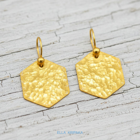 Hammered 24k gold, pure 9999 gold hexagon earrings with shiny forged gold textures on light grey old wooden surface, artist-jeweler 'Ella Kripaka' branding