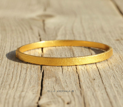 Hammered 24k 9999 gold bangle thick bracelet Ancient Rome style on a rustic light wooden background, branding artist-jeweler name 'Ella Kripaka'