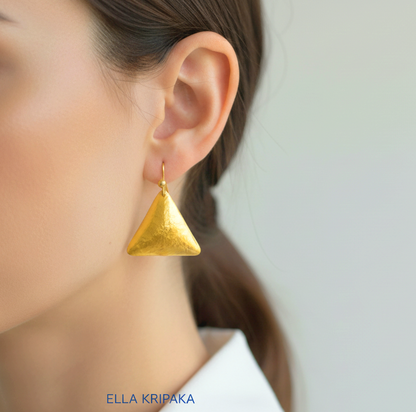 Hammered 24k gold, pure 9999 gold convex triangle earring with shiny forged gold textures worn on a woman and with light grey background, artist-jeweler 'Ella Kripaka' branding