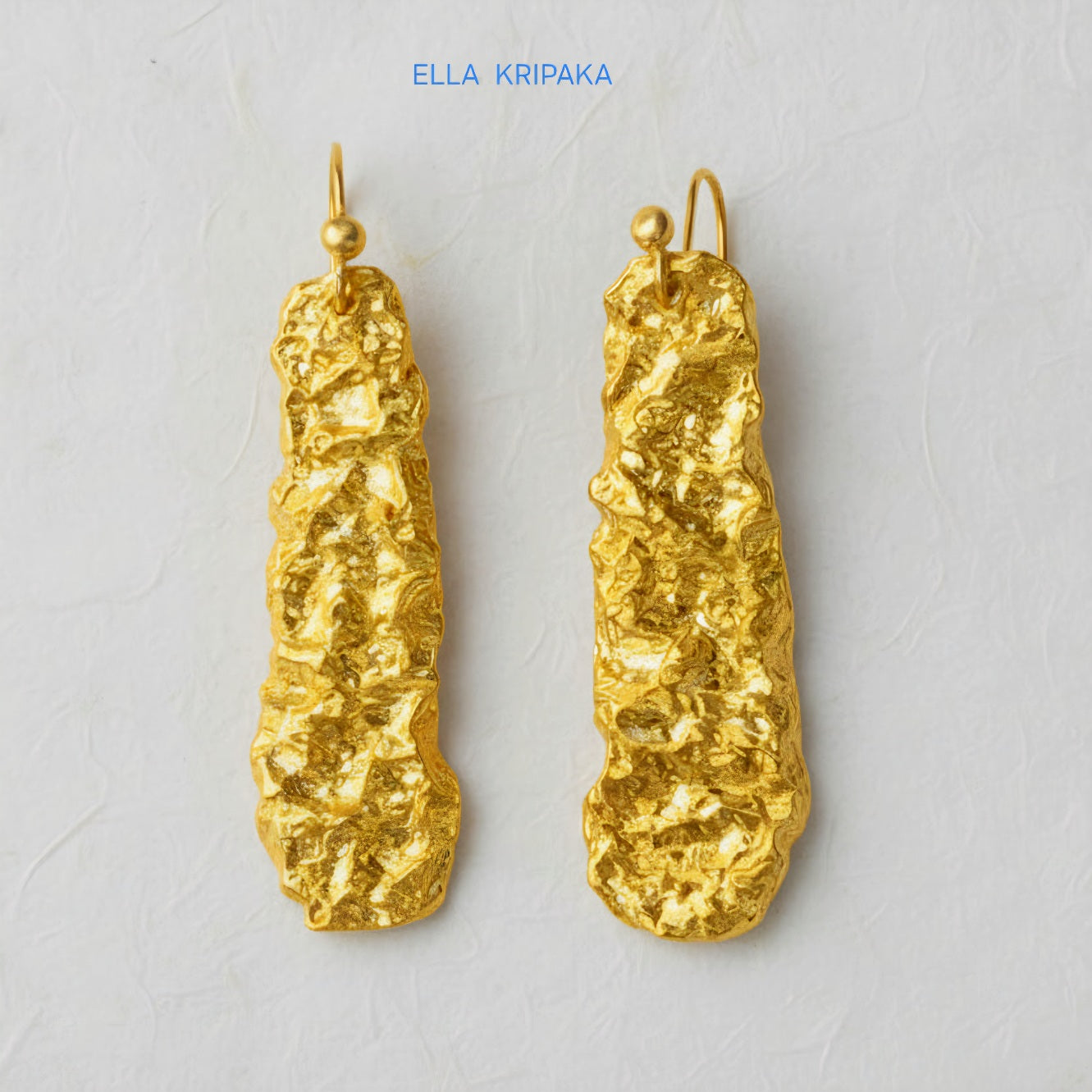 Organic hammered uneven 24k 9999 Gold earrings on a light grey surface, and with 'ELLA KRIPAKA' branding.