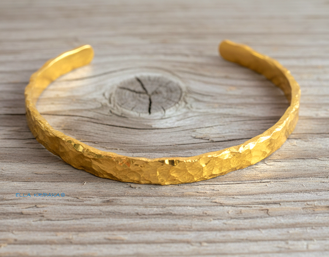 Organic hammered 24k gold bracelet cuff with uneven edges and textured design is on light wooden surface, branding artist's name 'Ella Kripaka'