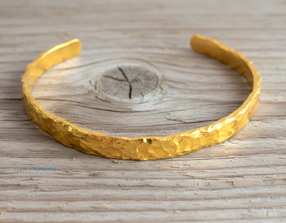 Organic hammered 24k gold bracelet cuff with uneven edges and textured design is on light wooden surface, branding artist's name 'Ella Kripaka'