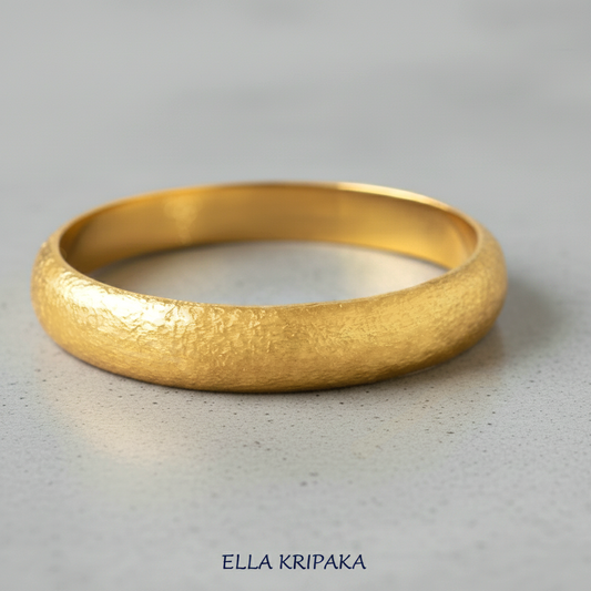 Hammered 24k gold bangle thick bracelet with rounded edges on grey polishing concrete background, and artist-jeweler 'Ella Kripaka' branding