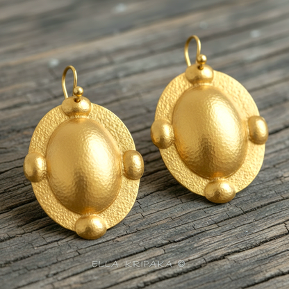 Hammered 24k gold, pure 9999 gold Byzantine convex ovals earrings with shiny forged gold textures on old wooden surface, artist-jeweler 'Ella Kripaka' branding