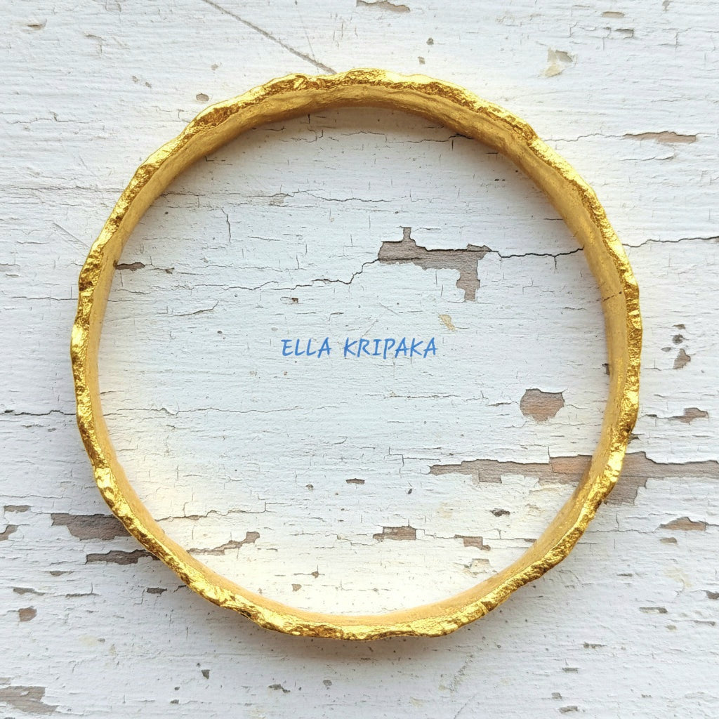 Hammered 22k gold organic textured bangle bracelet with uneven edges on old white wooden surface with 'Ella Kripaka' branding