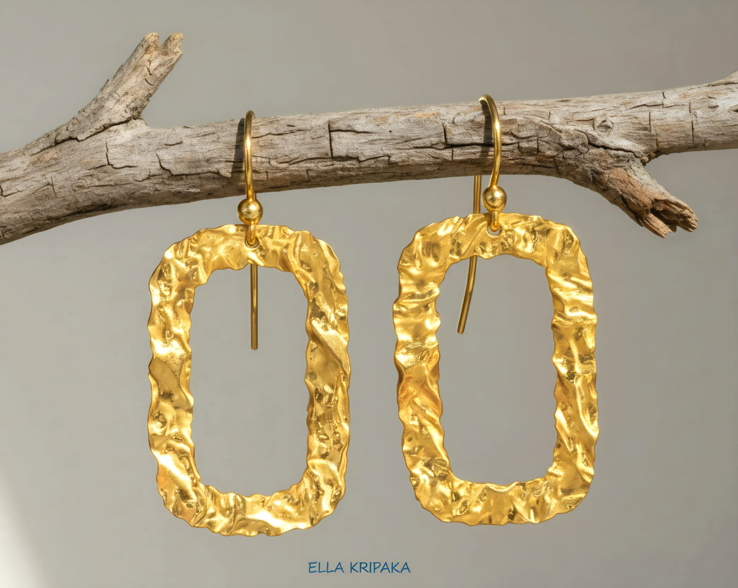 Organic hammered uneven 24k 9999 Gold hoops earrings hanging on a branch and on a grey background, and with 'ELLA KRIPAKA' branding
