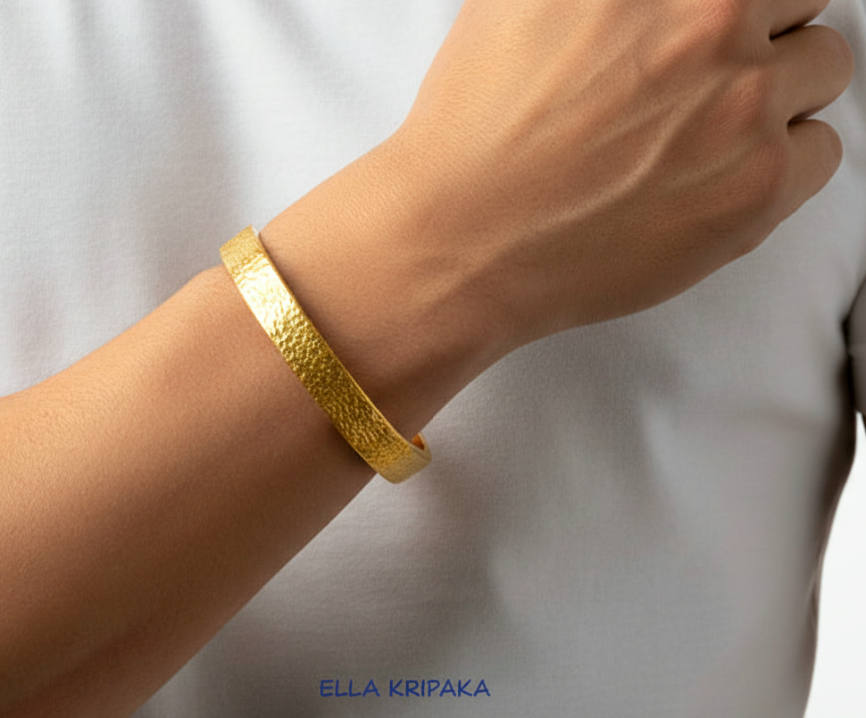 Hammered 24k Gold textured thick bangle bracelet worn by man on wrist and on blurred white t-shirt background, artist's name 'Ella Kripaka' branding