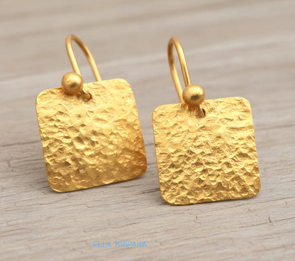 Hammered 24k gold, pure 9999 gold square earrings with shiny forged gold textures on old wooden surface, artist-jeweler 'Ella Kripaka' branding