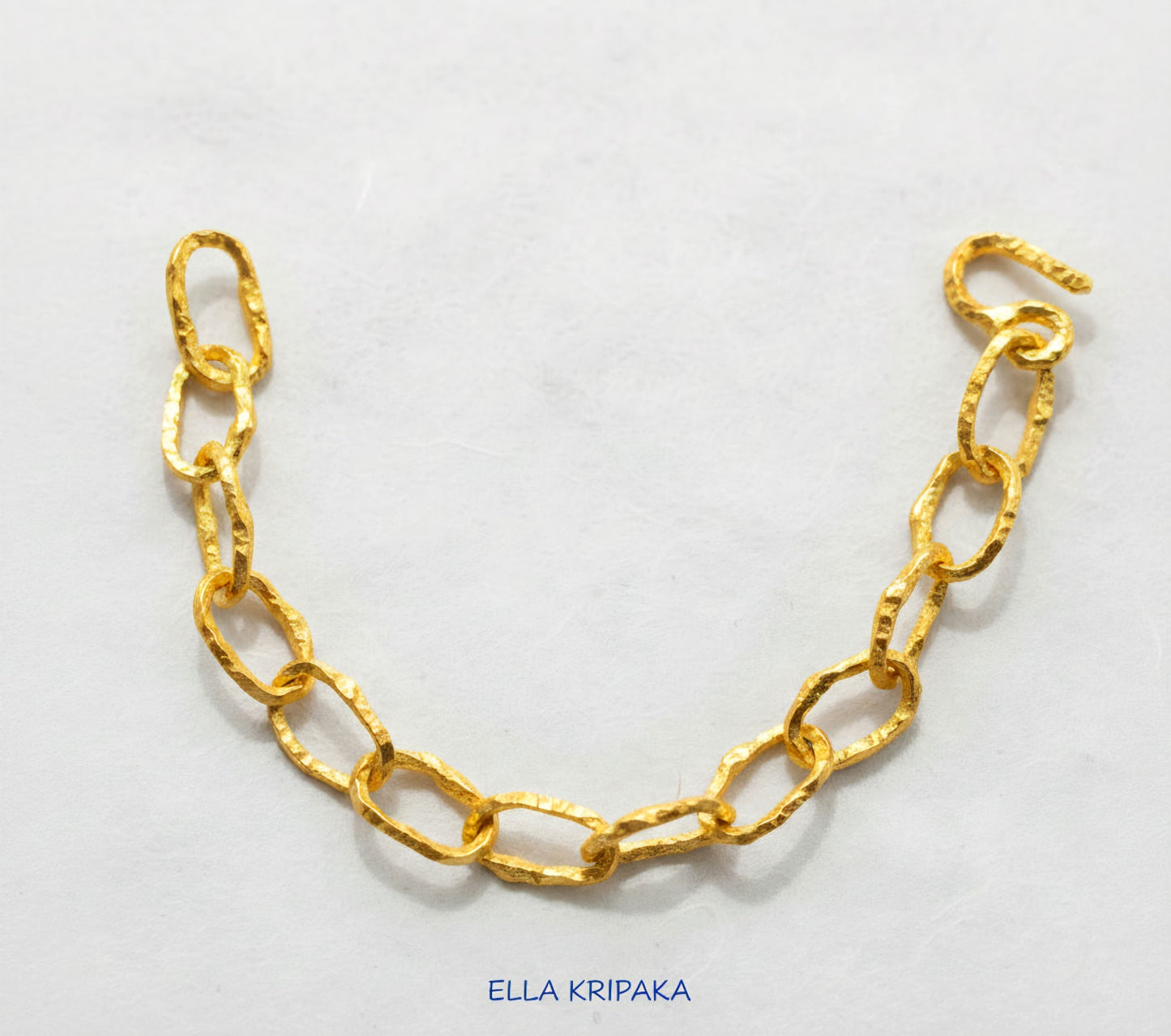 Hammered 24k 9999 gold organic bracelet chain with uneven textures and a thick hook clasp on white paper surface, artist name 'Ella Kripaka'