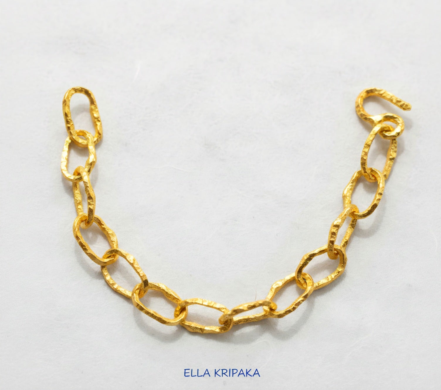 Hammered 24k 9999 gold organic bracelet chain with uneven textures and a thick hook clasp on white paper surface, artist name 'Ella Kripaka'