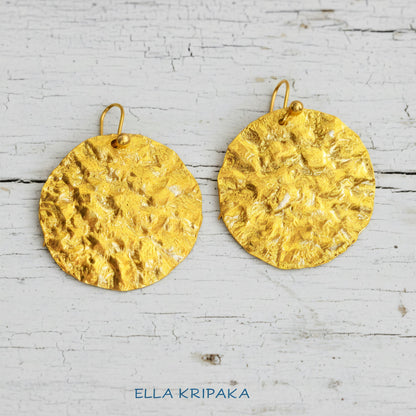 Organic 24k 9999 gold hammered wavy disk uneven earrings with shiny gold textures are on old white wooden background, artist-jeweler's name 'Ella Kripaka'