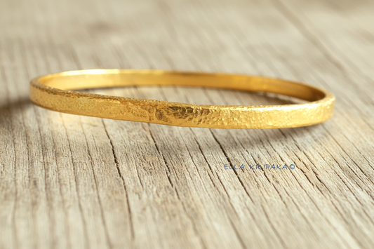Hammered 24k 9999 gold bangle thick bracelet Ancient Rome style on a wooden background, branding artist-jeweler name 'Ella Kripaka'