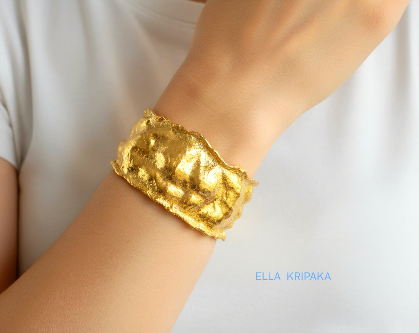 Organic 24k gold hammered bracelet cuff with a textured surface on a woman's wrist and with blurred light background, branded 'Ella Kripaka'