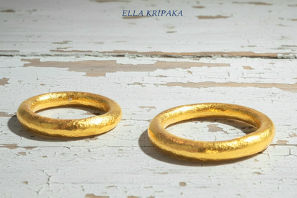 Two hammered 24k gold rounded textured rings on a white rustic wooden surface with 'Ella Kripaka' branding