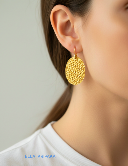 Organic 24k 9999 gold hammered oval earrings with shiny gold textures worn by a woman and on grey background, artist-jeweler's name 'Ella Kripaka'