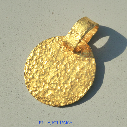 Hammered 24k gold, pure 9999 gold disk pendant with shiny forged gold textures on grey concrete surface, artist-jeweler 'Ella Kripaka' branding