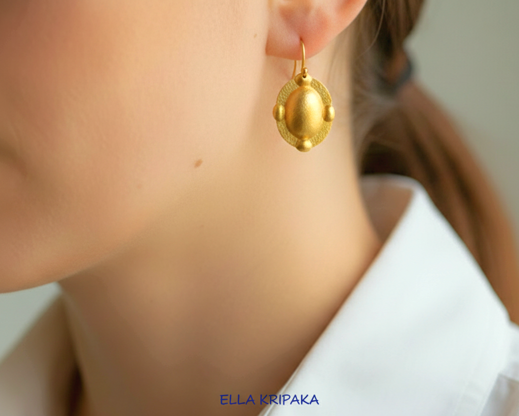 Hammered 24k gold, pure 9999 gold convex ovals Byzantine earring with shiny forged gold textures worn on a woman and with light grey background, artist-jeweler 'Ella Kripaka' branding