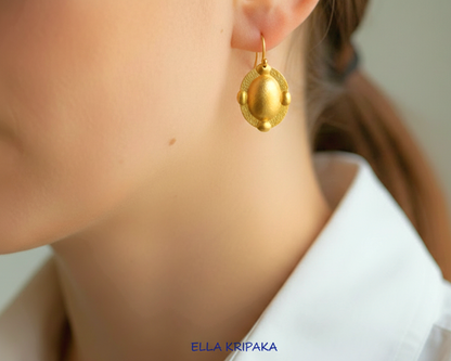 Hammered 24k gold, pure 9999 gold convex ovals Byzantine earring with shiny forged gold textures worn on a woman and with light grey background, artist-jeweler 'Ella Kripaka' branding