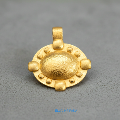 Hammered 24k gold Byzantine convex pendant on a grey concrete surface with 'Ella Kripaka' branding