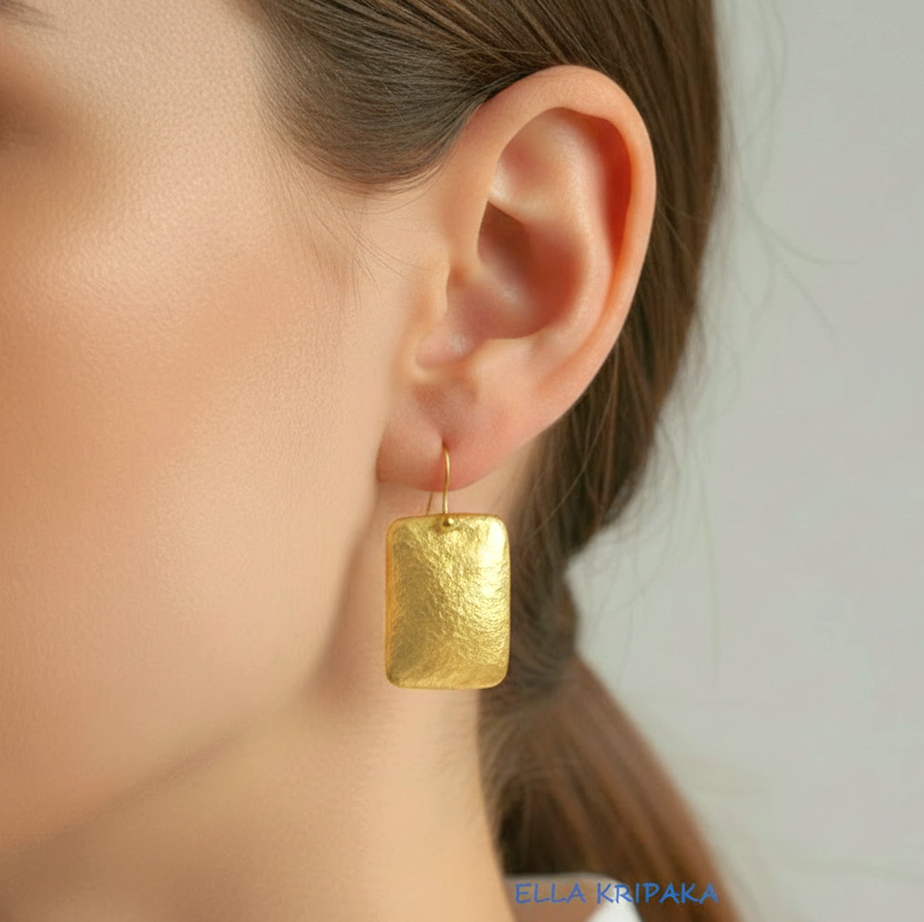 Hammered 24k gold, pure 9999 gold convex rectangular earring with shiny forged gold textures worn on a woman and with light grey background, artist-jeweler 'Ella Kripaka' branding