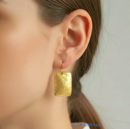 Hammered 24k gold, pure 9999 gold convex rectangular earring with shiny forged gold textures worn on a woman and with light grey background, artist-jeweler 'Ella Kripaka' branding