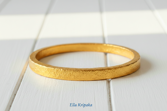 Hammered 24k 9999 gold bangle thick bracelet Ancient Rome style on a white wooden background, artist-jeweler name 'Ella Kripaka' branding