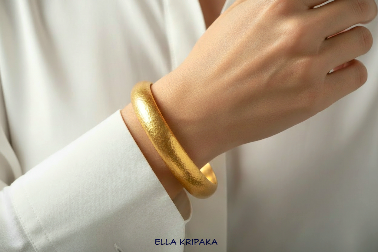Hammered 24k gold bangle thick bracelet with rounded edges is worn by a woman on wrist and on white shirt background, artist-jeweler 'Ella Kripaka' branding