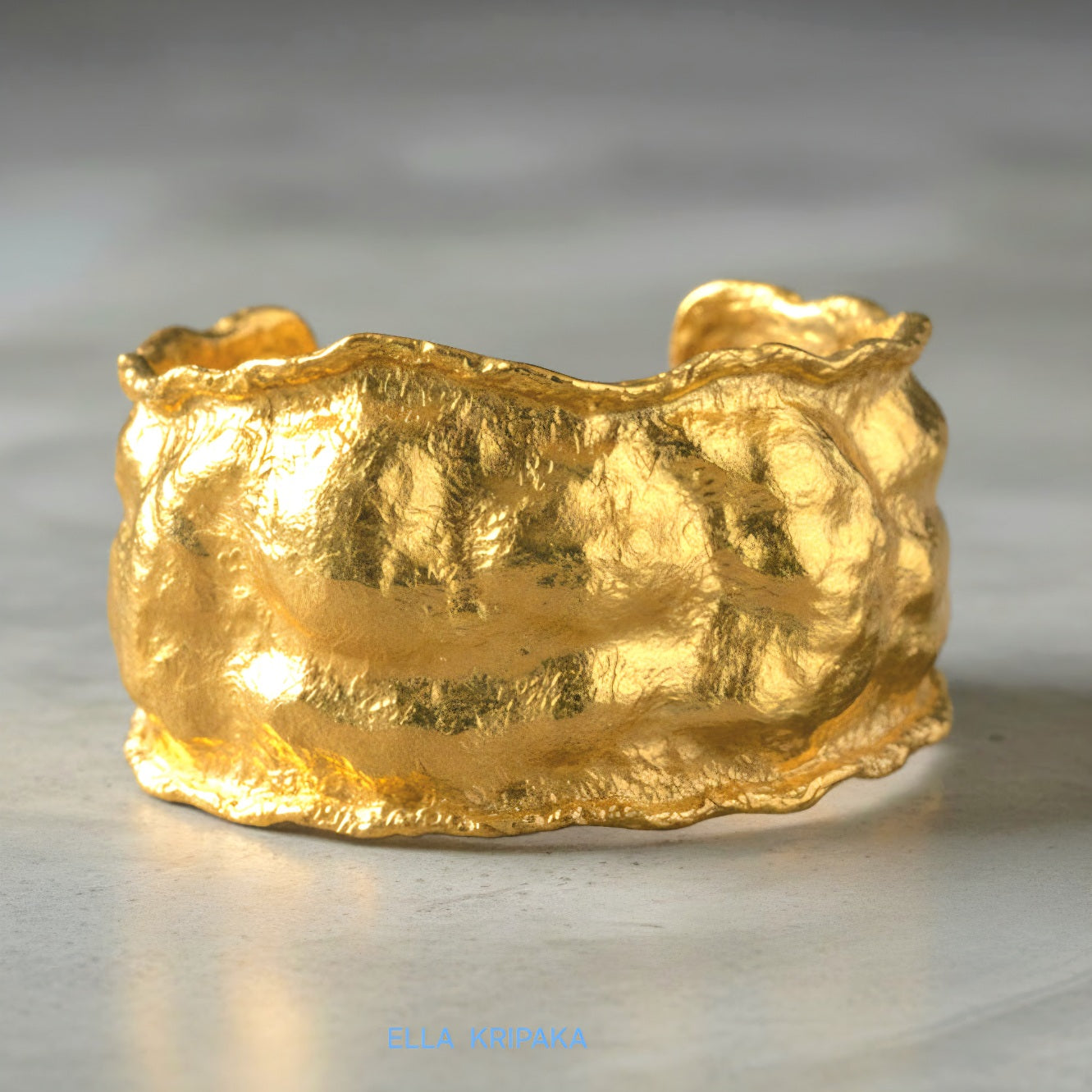 Organic 24k gold hammered bracelet cuff with a textured surface on a grey concrete background, branded 'Ella Kripaka'