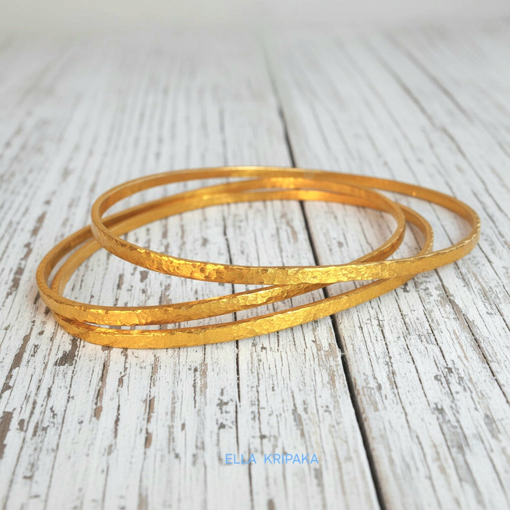 Hammered 22k gold three textured bangles bracelets Ancient Rome style on old white wooden surface with 'Ella Kripaka' branding