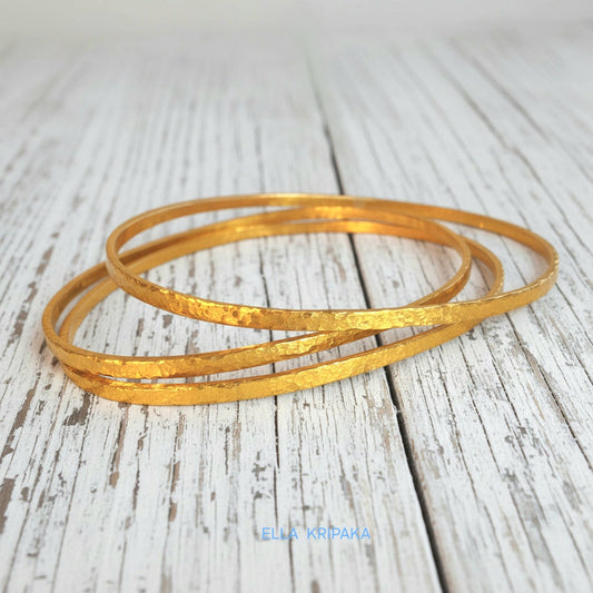 Hammered 22k gold three textured bangles bracelets Ancient Rome style on old white wooden surface with 'Ella Kripaka' branding